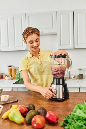 happy woman preparing delicious smoothie while grinding fresh fruits in kitchen, vegetarian recipe