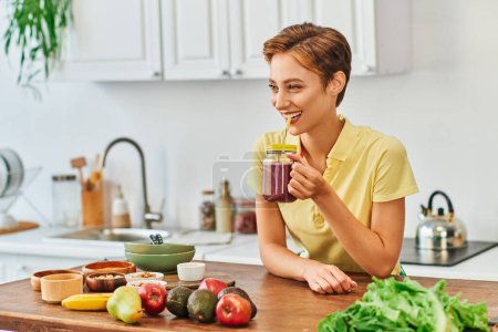 smiling vegetarian woman drinking delicious smoothie from mason jar with straw in kitchen