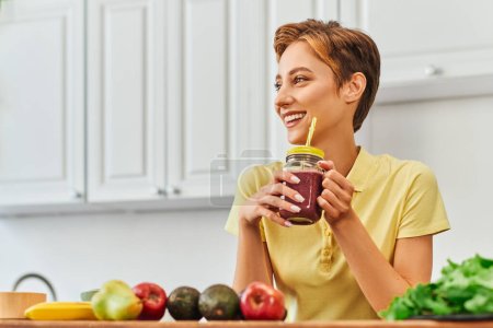 fröhliche Vegetarierin hält frischen Smoothie in Einmachglas mit Stroh und schaut in Küche weg