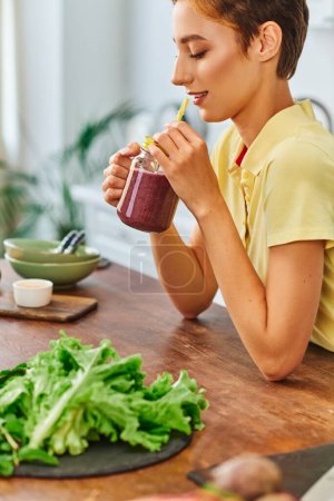 Photo for Young vegetarian woman holding mason jar with delicious smoothie near fresh lettuce on kitchen table - Royalty Free Image