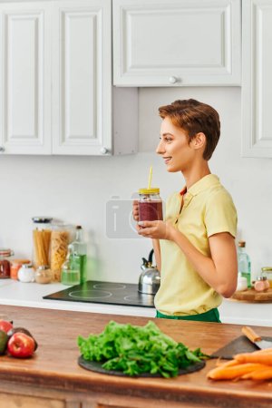 Photo for Side view of smiley vegetarian woman holding mason jar with fresh smoothie in modern kitchen - Royalty Free Image