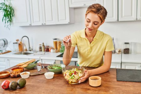 smiling woman seasoning fresh fruit salad with sesame seeds in kitchen, delicious vegetarian diet