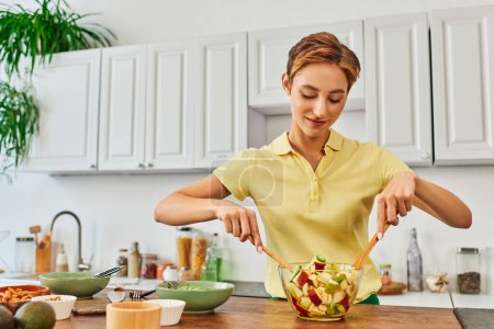 Photo for Young smiling woman mixing delicious fruit salad while cooking in kitchen, plant-based diet concept - Royalty Free Image
