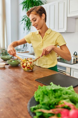 Photo for Young and joyful woman mixing fresh fruit salad while cooking vegetarian meal in modern kitchen - Royalty Free Image