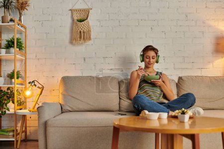 delighted woman in headphones having vegetarian dinner on couch in cozy living room, quiet evening