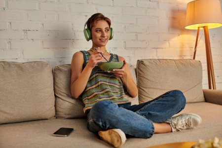 smiling woman in headphones with salad bowl on couch in living room, vegetarian evening snack