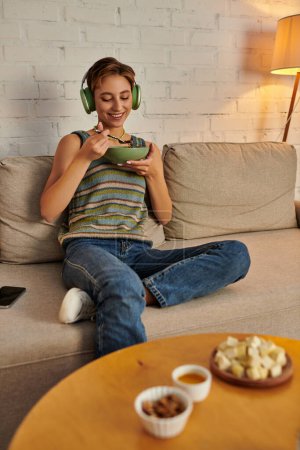 fröhliche Frau mit Kopfhörer beim vegetarischen Abendessen in der Nähe auf der Couch am Tisch mit pflanzlicher Mahlzeit