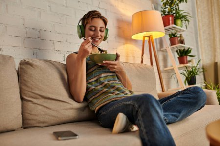 fröhliche Frau mit Kopfhörer beim vegetarischen Abendsnack auf der Couch im gemütlichen Wohnzimmer