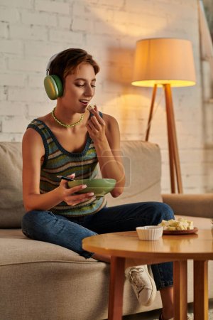 mujer vegetariana en auriculares sosteniendo ensaladera y comiendo queso de tofu, merienda por la noche en el sofá