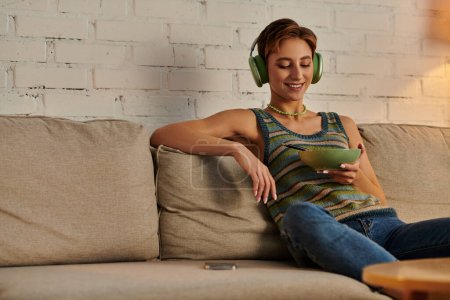 mujer veggie complacido en los auriculares sentado con ensaladera en el sofá en la acogedora sala de estar por la noche