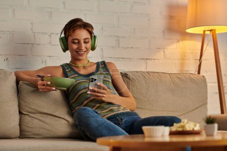 mujer veggie feliz en la red de auriculares en el teléfono inteligente mientras está sentado con ensaladera en el sofá