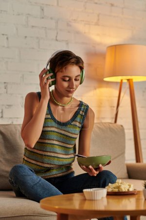 mujer vegetariana con ensaladera escuchando música en auriculares cerca de queso de tofu en la sala de estar