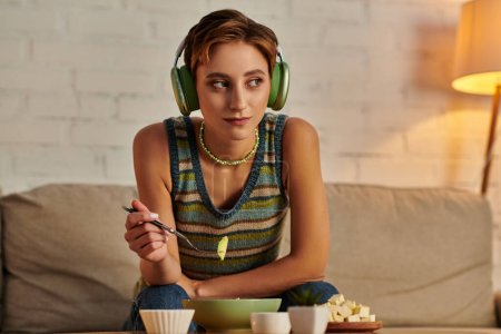 mujer de ensueño en auriculares mirando hacia otro lado mientras toma merienda vegetariana por la noche en el sofá en casa