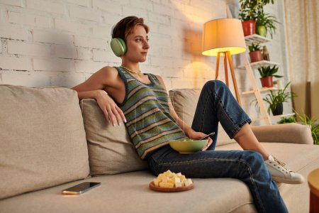 verträumte Gemüsefrau mit Kopfhörer sitzt mit Salatschüssel neben Tofu-Käse und Smartphone auf der Couch