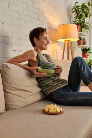 Lächelnde Frau mit Salatschüssel auf Couch im Wohnzimmer in der Nähe von Tofu-Käse, vegetarisches Abendessen