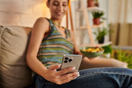 blurred vegetarian woman taking selfie while sitting with salad bowl on couch in living room