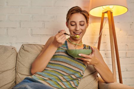 Zufriedene junge Frau isst leckeren vegetarischen Salat beim abendlichen Imbiss im gemütlichen Wohnzimmer