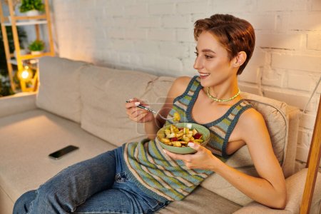 Lächelnde Frau sitzt mit Schüssel leckerem vegetarischem Obstsalat auf Couch im Wohnzimmer