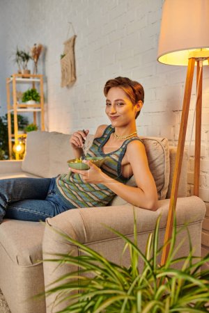 young vegetarian woman smiling at camera while eating fruit salad on sofa in cozy living room