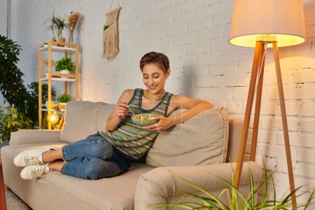 entzückte Gemüsefrau sitzt auf Couch neben Lampe und isst frischen Obstsalat auf gemütlichem Sofa