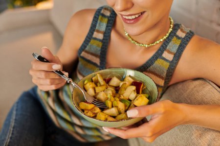 abgeschnittene Ansicht einer lächelnden Frau mit einer Schüssel leckerem Obstsalat auf dem heimischen Sofa, pflanzliche Ernährung