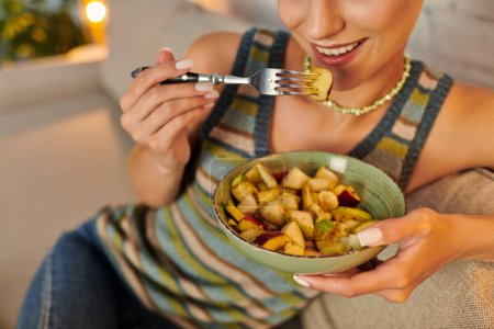 abgeschnittene Ansicht einer vegetarischen Frau, die köstlichen Obstsalat zum Abendessen auf dem Sofa im Wohnzimmer isst