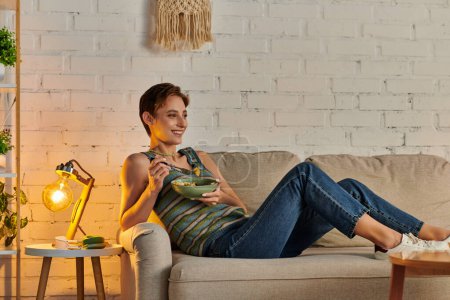 glückliche Frau mit Schüssel vegetarischem Salat auf Sofa im Wohnzimmer neben moderner Schreibtischlampe