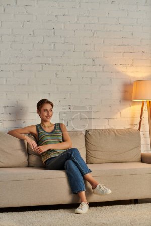 glückliche junge Frau sitzt auf gemütlicher Couch neben Lampe im modernen Wohnzimmer, Freizeit am ruhigen Abend