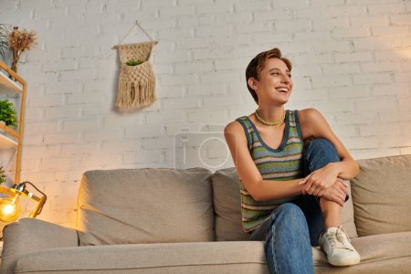 Foto de Joven mujer emocionada mirando hacia otro lado en el sofá acogedor en la sala de estar moderna, feliz vida en casa - Imagen libre de derechos