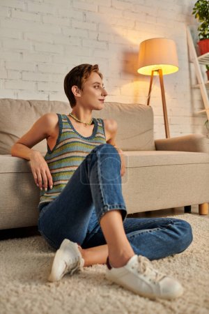 positive and dreamy young woman looking away while sitting on floor near couch in living room