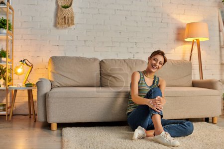 glückliche junge Frau sitzt auf dem Boden neben Couch im modernen Wohnzimmer und lächelt abends in die Kamera