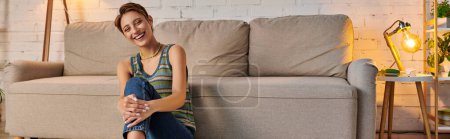 joyous young woman sitting near couch in cozy living room and looking at camera, horizontal banner
