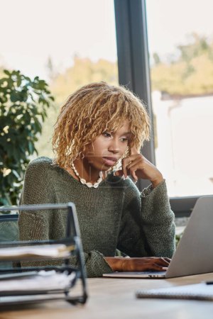 focused young african american woman with curly hair looking at her laptop, working process
