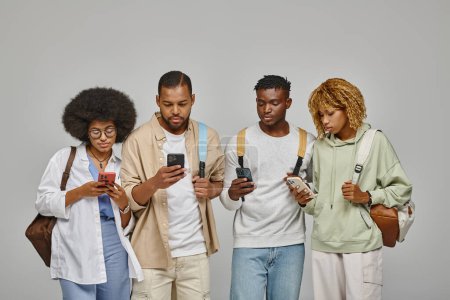 Foto de Grupo de amigos en atuendos casuales con mochilas mirando sus teléfonos móviles, estudiando concepto - Imagen libre de derechos