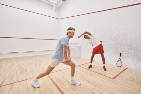 young redhead man with headband doing lunges near african american friend inside squash court