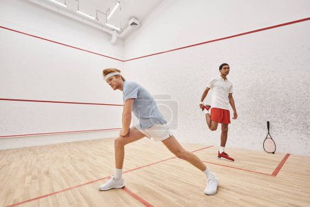 sporty redhead man with headband doing lunges near african american friend inside squash court