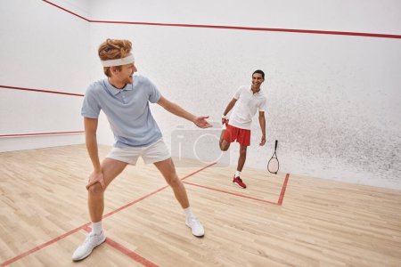 happy redhead man with headband doing lunges near african american friend inside squash court