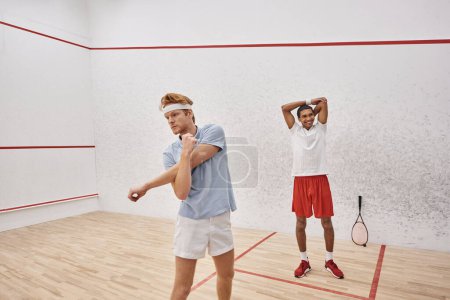 young redhead man with headband warming up near happy african american friend inside squash court