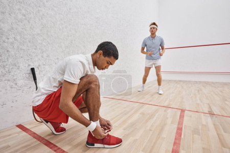 african american man tying shoelaces while sitting inside of squash court near redhead friend