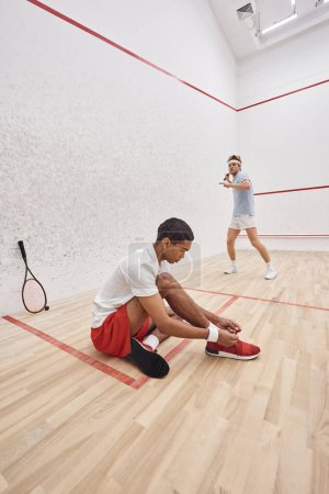 african american man tying shoelaces while sitting near redhead friend inside of squash court