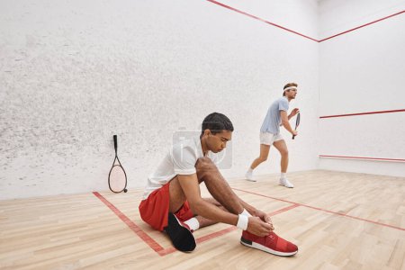 african american player tying shoelaces while sitting near redhead friend inside of squash court
