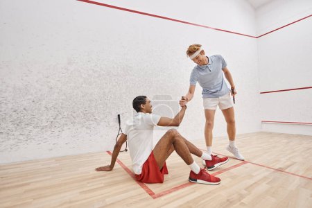 happy redhead man helping his african american friend to stand up from floor inside of squash court