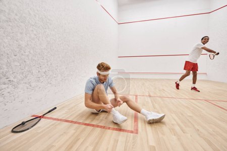 redhead man tying shoelaces while sitting near african american friend inside of squash court