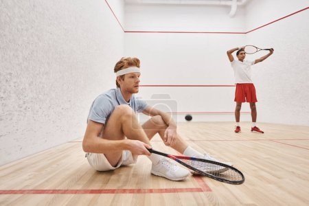 man in sportswear holding racquet while sitting near african american friend inside of squash court