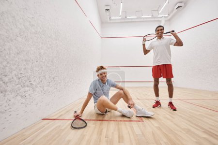 happy man holding racquet while sitting near african american friend inside of squash court