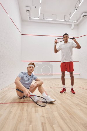 happy man holding racquet while sitting near african american player inside of squash court