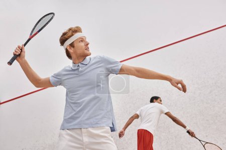 redhead man in sportswear and headband playing with african american friend inside of squash court
