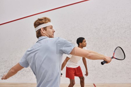 redhead man in sportswear and headband playing with african american friend inside of squash court