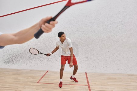 african american man in sportswear playing with friend inside of squash court, blurred foreground
