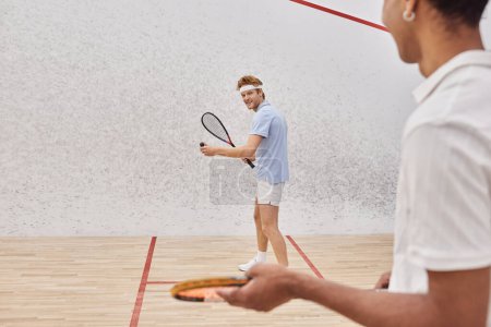 focus on happy man in sportswear playing with african american friend inside of squash court